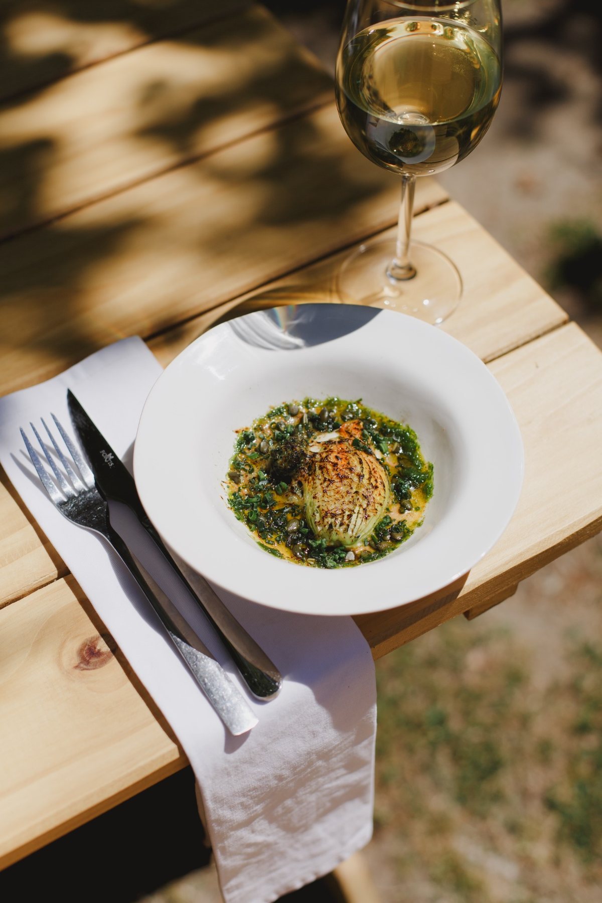 A seasonal dish with green herbs in a white bowl, beside a glass of wine